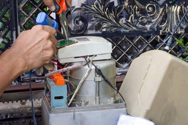 A person is using a screwdriver and pliers on an exposed electronic device next to an ornate black metal driveway gate. Wires and circuit boards are visible, with a beige plastic cover partially seen on the right, suggesting some gate repair work is underway.