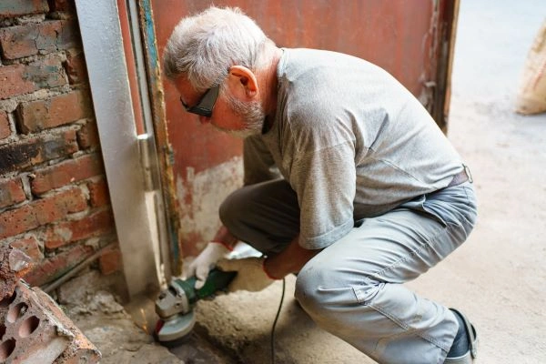 A man wearing safety glasses and gloves crouches while using an angle grinder to cut or polish a metal beam near a brick wall. He appears to be performing construction or repair work, possibly installing components for an electric gate.
