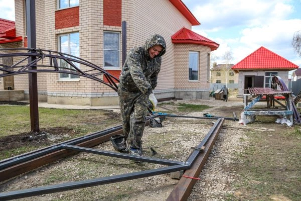 A gate technician in camouflage clothing works on the metal framework outside a brick house with red roofs. The person uses a tool while standing among various construction materials, and a second building with a similar red roof is visible in the background, possibly related to a driveway gate or gate opener installation.