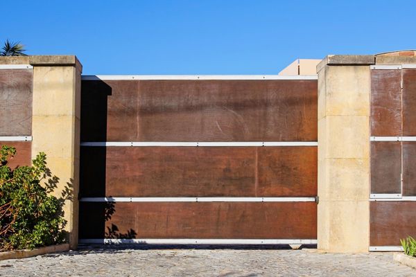 A large, closed wooden gate with metal reinforcements is flanked by two stone pillars. The gate, reminiscent of local craftsmanship in Dallas, is part of a fence that runs off the sides of the image. A clear blue sky is in the background and greenery is partially visible on the left.