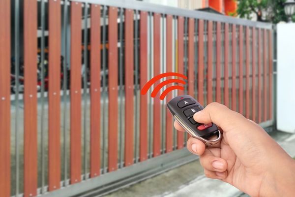 A person is using a remote control to operate an automatic sliding gate made of vertical wooden slats with metal frames. The red signal waves indicate the remote is active and operating the gate, which was recently repaired by a Dallas company specializing in gate maintenance.