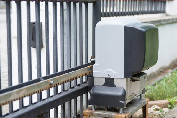 An automated gate motor is installed on the ground next to a black metal gate. It is housed in a gray and black casing and connected to a rack and pinion system, enabling the local Dallas gate to open and close automatically. The background features a paved area and some greenery.