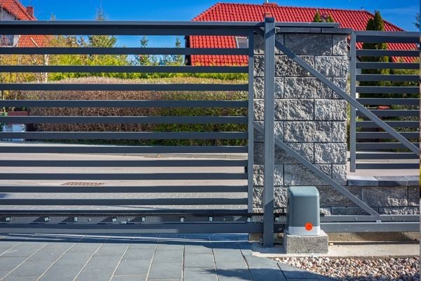 A modern, metal sliding gate stands in front of a Dallas residential property with a red-roofed house in the background. The gate, from a reputable company, is mounted on a stone pillar and slides along a ground track, with its control box visible at the base under the clear blue sky.