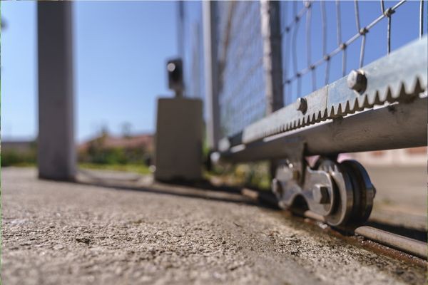 Close-up of a sliding gate wheel mechanism on a concrete driveway. The metal gate is mounted on a track, with a focus on the wheel and lower part of the gate. The background is slightly blurred, showing some greenery and a fence post, hinting at the need for occasional repair.