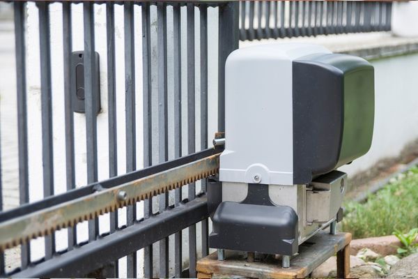 Close-up of an automatic sliding gate motor attached to a black iron gate at the end of a driveway. The motor, encased in black and gray housing, is mounted on a metal base. Rust patches are visible on the metal track near the motor. Grass and pavement surround the gate, indicating it may need repair soon.