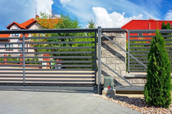 A modern, horizontal metal and stone gate entrance to a residential property in Dallas with red-roofed houses and greenery visible in the background. The pavement is smooth and well-kept, and a neatly trimmed bush is located beside the gate, reflecting local charm.