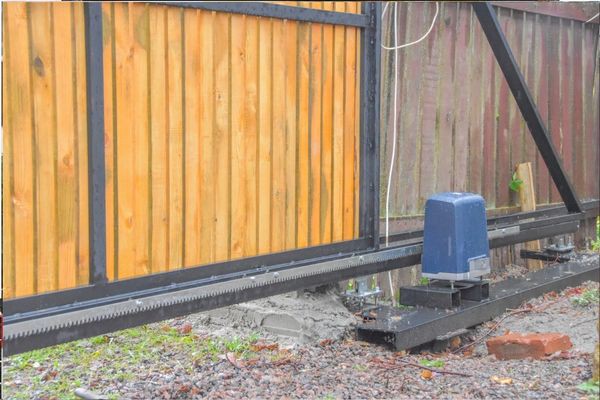 A sliding wooden gate with black metal frames and a motorized mechanism for automatic opening and closing, provided by a local company. The gate is partially open, exposing the motor unit fixed to the ground on a gravel surface. A brick and some debris are also visible nearby.