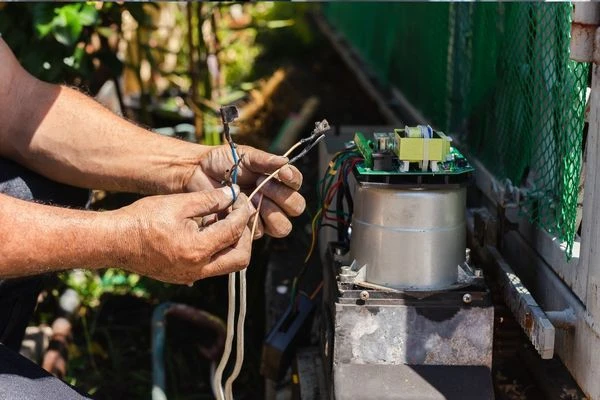 A person is working on electrical wires, holding tangled cables next to an exposed motor or generator with circuitry, outdoors. The repair work is being done beside a green mesh fence, and some plants are visible in the background, likely near a company's driveway.