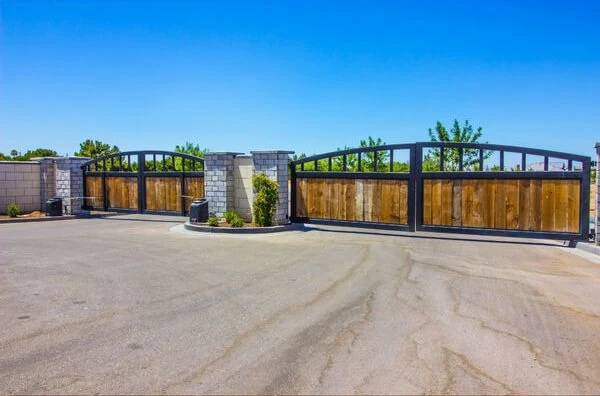 A pair of large, wooden, gated entrances with black metal frames stand open on a paved road in Dallas. The gates are flanked by stone pillars, with small plants and trees visible in the background under a clear blue sky.