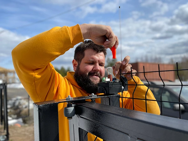 A local man with a beard wearing a yellow hoodie uses a screwdriver to repair a mechanism on a black metal gate under a partly cloudy sky in Dallas. He is working outdoors next to a wire fence.