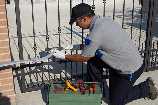 A local man in a grey and blue uniform kneels on the ground and repairs a metal gate using tools from a green toolbox placed nearby. The black gate, adorned with vertical bars and decorative elements, is being meticulously fixed by the Dallas resident, who wears gloves and a black cap.