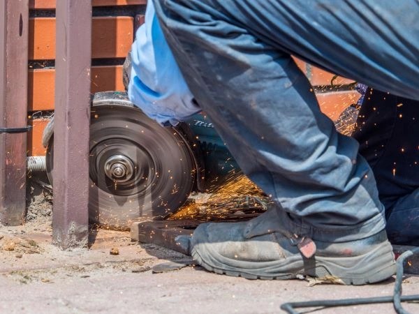 A person in blue work clothes and black protective shoes uses an angle grinder to cut through metal, with orange sparks flying. The individual is crouched near a brick wall and local company gate, with safety gear like gloves and a face shield partially visible.