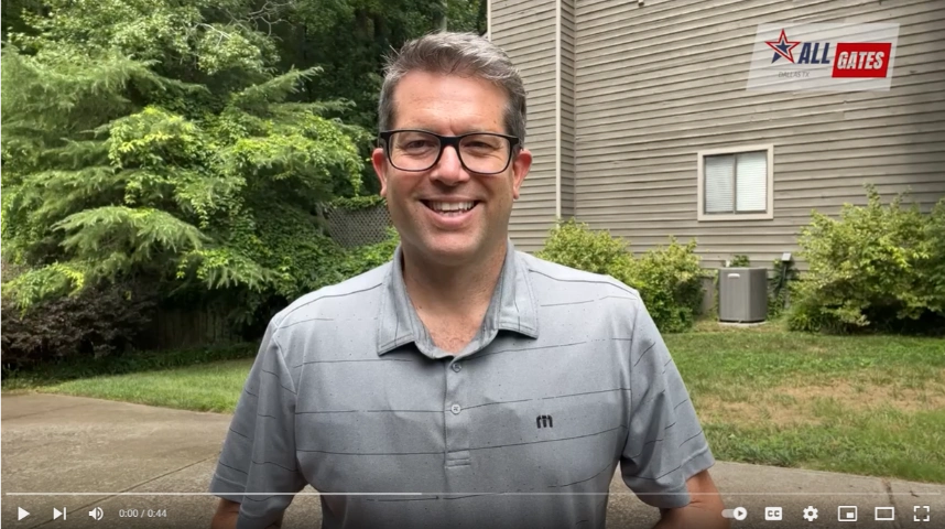 A person with short hair and glasses, wearing a gray collared shirt, is standing outside in front of a house. The house has horizontal siding and an air conditioning unit. The yard is green with trees and shrubbery, showcasing a well-maintained gate. The "All Gates Media" logo appears in the upper right corner.